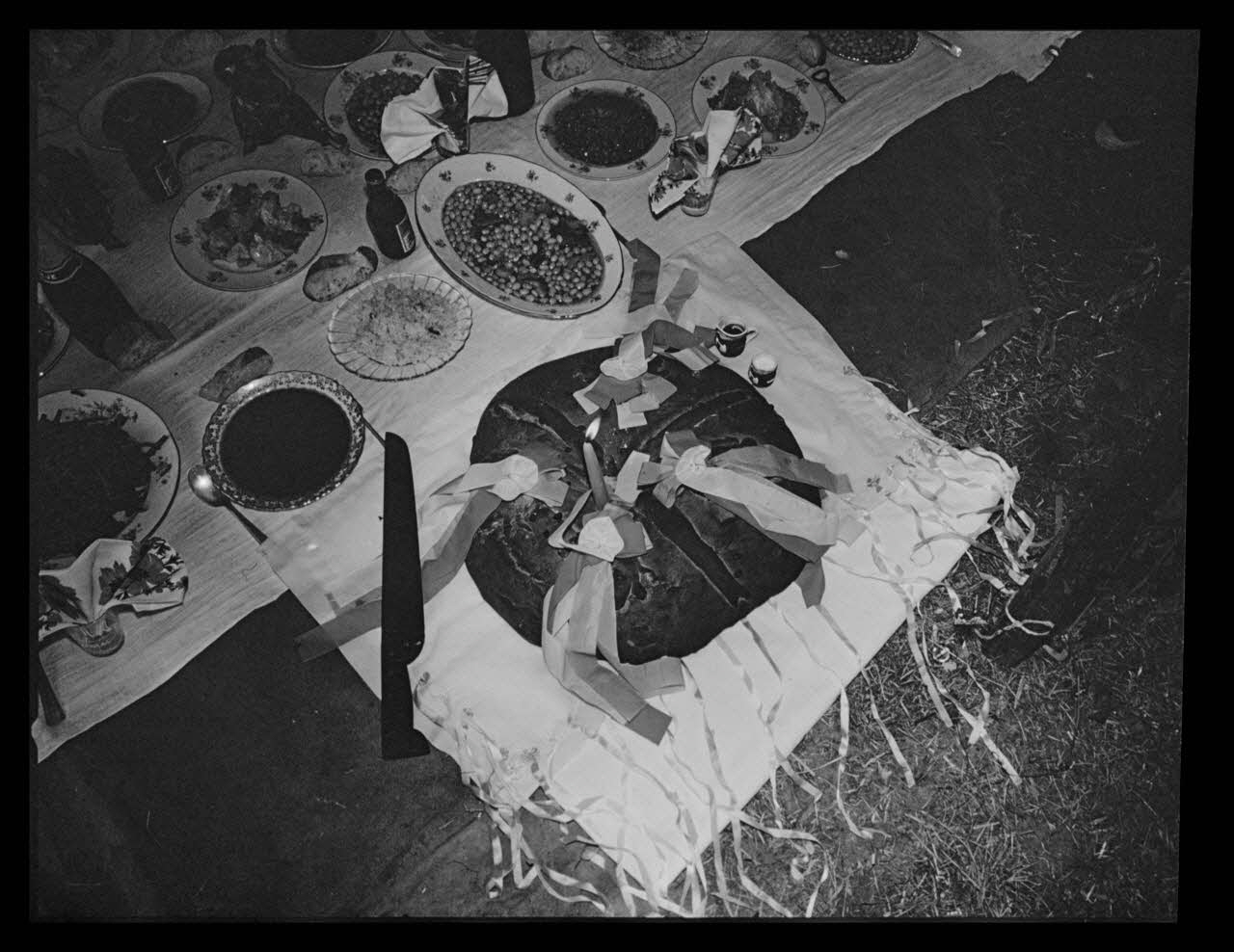 Philippe Lemaire De Marne photographie Roms, nomades "Kalderash". Fête traditionnelle "Prasmiko". Famille de "Papa" Gusman et Théodore Gusman, son frère dans un terrain loué à la sortie du village. Gâteau "Kolako" avec un couteau, une bougie allumée, un pot de vin et du sel Midi-Pyrénées, France 1965/8/15 Ph.1966.68.71 Photo