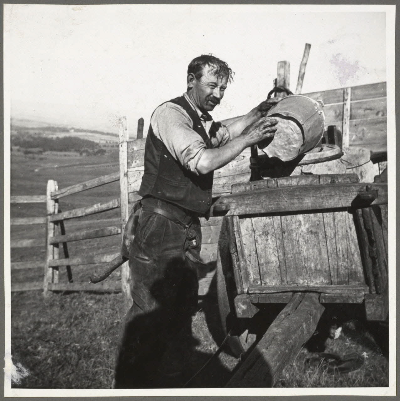 Anonyme photographie Vacher avec sa selle, siège à un seul pied. Dans le Cantal, sur les hauts pâturages de montagne où les vacheries passent des mois d'estive, le vacher a souvent un grand nombre de bêtes à traire. S'il utilisait un petit banc, comme cela se fait ailleurs, il n'aurait pas ses deux mains libres et perdrait du temps pour le déplacer. Dans la Haute-Auvergne, le vacher se sert d'un siège à un seul pied - la selle - fixé à demeure à son postérieur pendant toute la durée de la traite, à l'aide de deux courroies de cuir passant entre les jambes et venant se rattacher solidement à la selle à la hauteur des reins. Le vacher a ainsi ses mains disponibles. Quand il s'accroupit, la selle forme avec ses jambes un trépied sur lequel le buste repose d'une façon très stable. A noter que les bergers se plaisent à donner souvent à la selle une forme phallique qui leur fait une allure étrange quand ils sont debout. (Henri Lelong) Auvergne, France 1937 Ph.1937.947.B Photo