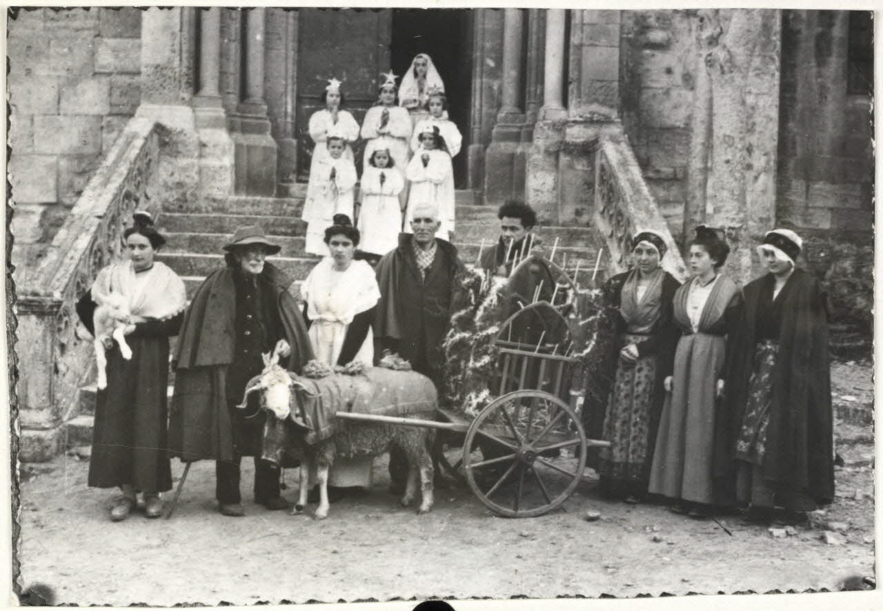 Anonyme photographie Noël aux Baux. La "charrette de l'agneau", bergers, femmes et enfants devant l'église. Exemple de pastrage. La charrette trainée par un floucat avec collier et redon transporte jusqu'à l'église un jeune agneau. Celui-ci sera amené en offrande à leur tour. Ici l'agneau est dans les bras d'une femme habillée en arlésienne (tout à fait à gauche) et les participants du pastrage dont deux bergers avec leur ample cape à pélerine caractristique des bergers provençaux posent pour le photographe devant l'église des Baux" Provence-Alpes-Côte d'Azur, France 1950 Ph.1962.131.1 Photo