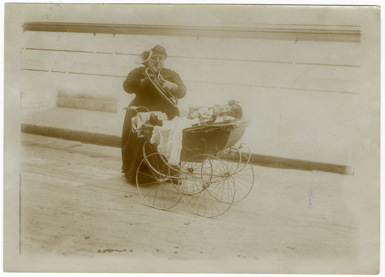 Branger (Paris) photographie Le 14 juillet à Paris. Pour soulager sa misère, une pauvre mère poussant dans une petite voiture une petite fille infirme, implore la pitié des promeneurs en jouant de la basse Ile-de-France, France 1914/7/14 1997.27.3.10 Photo